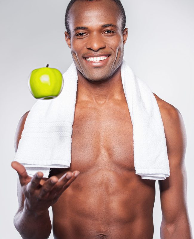 Enjoying healthy lifestyle. Young muscular African man with towel on shoulder throwing up an apple and smiling at camera while standing against grey background